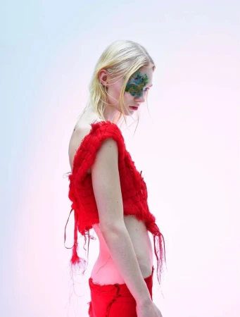 Blonde fashion model in red knit top and pants poses in a side profile against a light gradient background. Face paint detail.