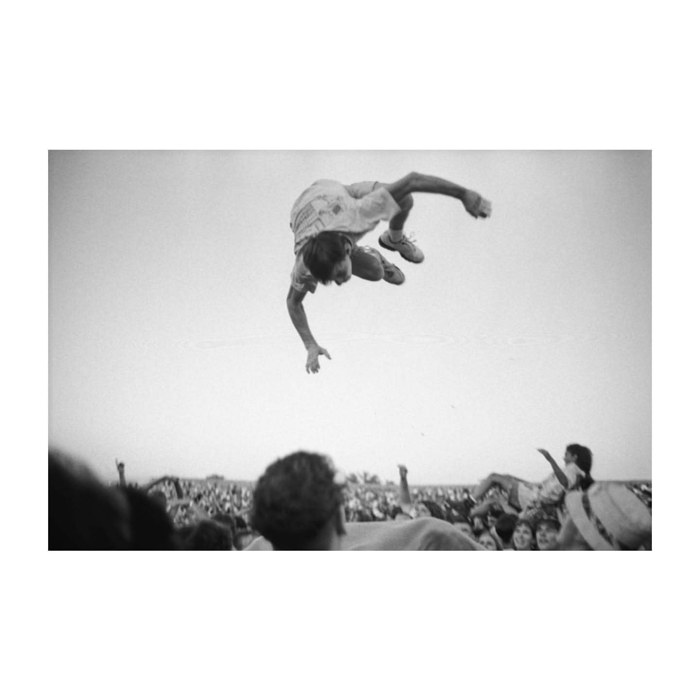 A person in mid-air performing a flip with a large crowd visible in the background under a bright sky in a monochrome image.