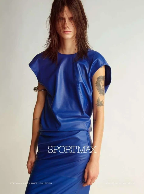 Fashion shot of a woman in a bright blue skirt and top with wet hair, arm jewelry and a lion tattoo in a neutral studio setting.