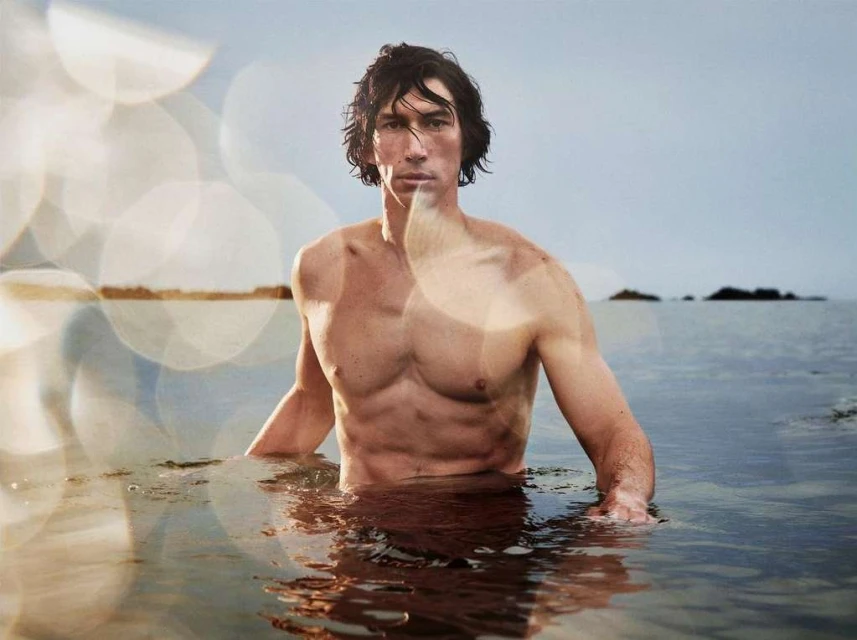 Shirtless Adam Driver stands in ocean water with bokeh foreground. Wet dark hair, light skin, looking at camera, body partially submerged.
