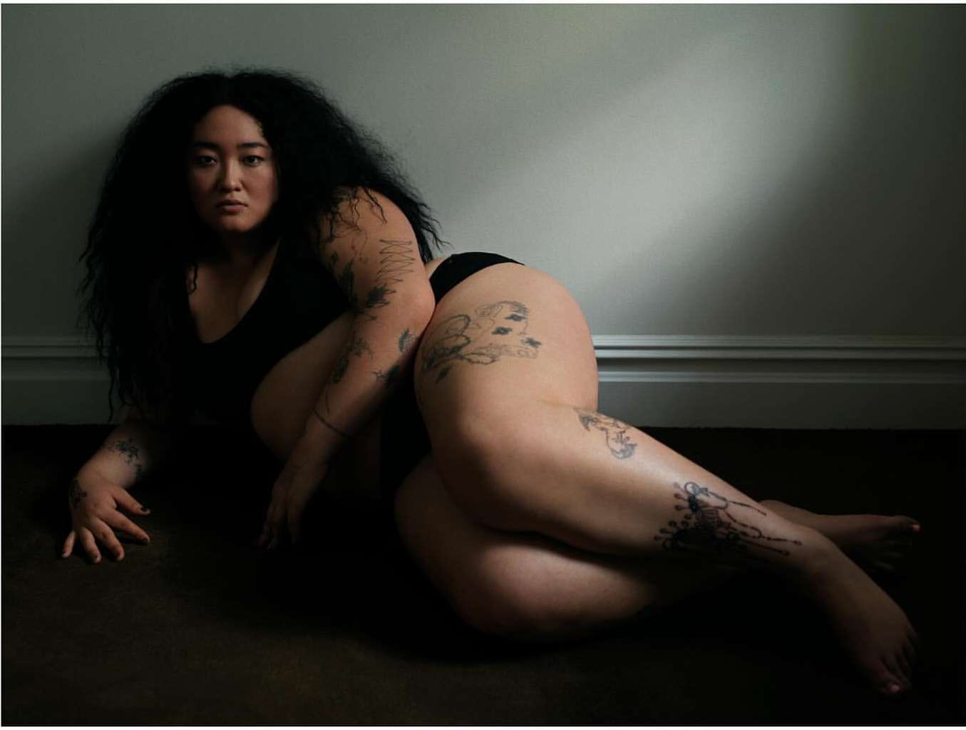 Portrait of a person with dark hair, tattoos, laying on the floor, wearing black undergarments against a grey wall. Indoor setting.