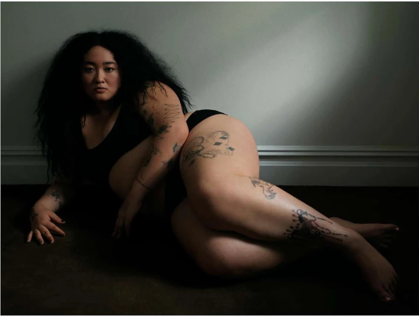 Portrait of a person with dark hair, tattoos, laying on the floor, wearing black undergarments against a grey wall. Indoor setting.