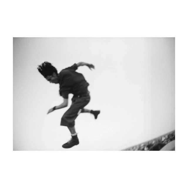 Monochrome shot of a man in mid-air, appearing to jump or leap, with his body angled and hair flying, set against a bright sky.