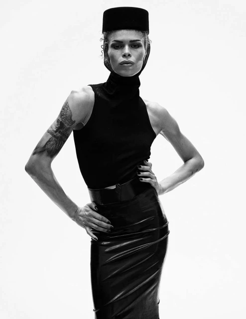 Black and white portrait of a stylish adult fashion model in a black turtleneck, pencil skirt, and hat against a white backdrop.