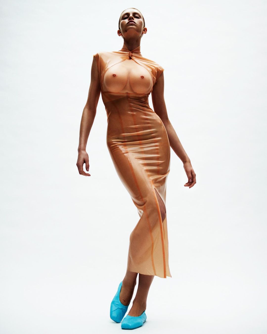 Woman in peach latex dress and blue flats poses in a fashion shot against a white background in a studio with bright lighting.