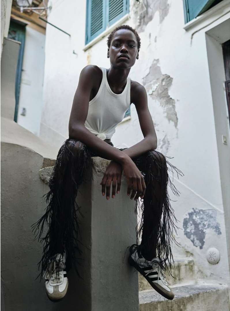 A slender African American woman in a white tank top and black fringe pants sits on a stone step in an urban outdoor setting.