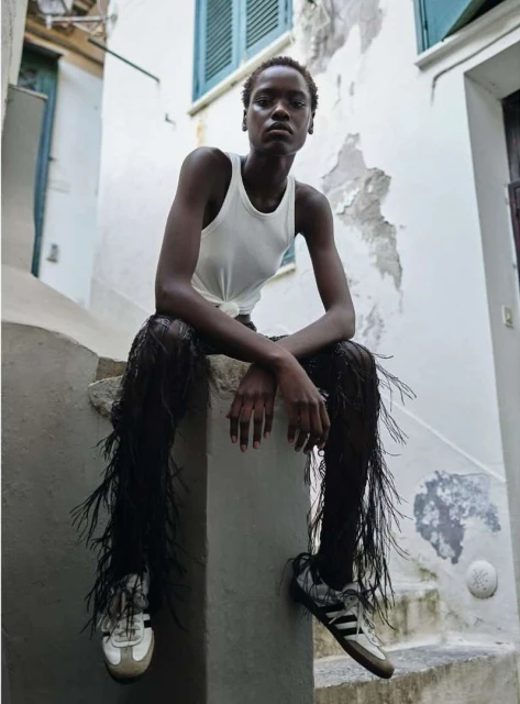 A slender African American woman in a white tank top and black fringe pants sits on a stone step in an urban outdoor setting.