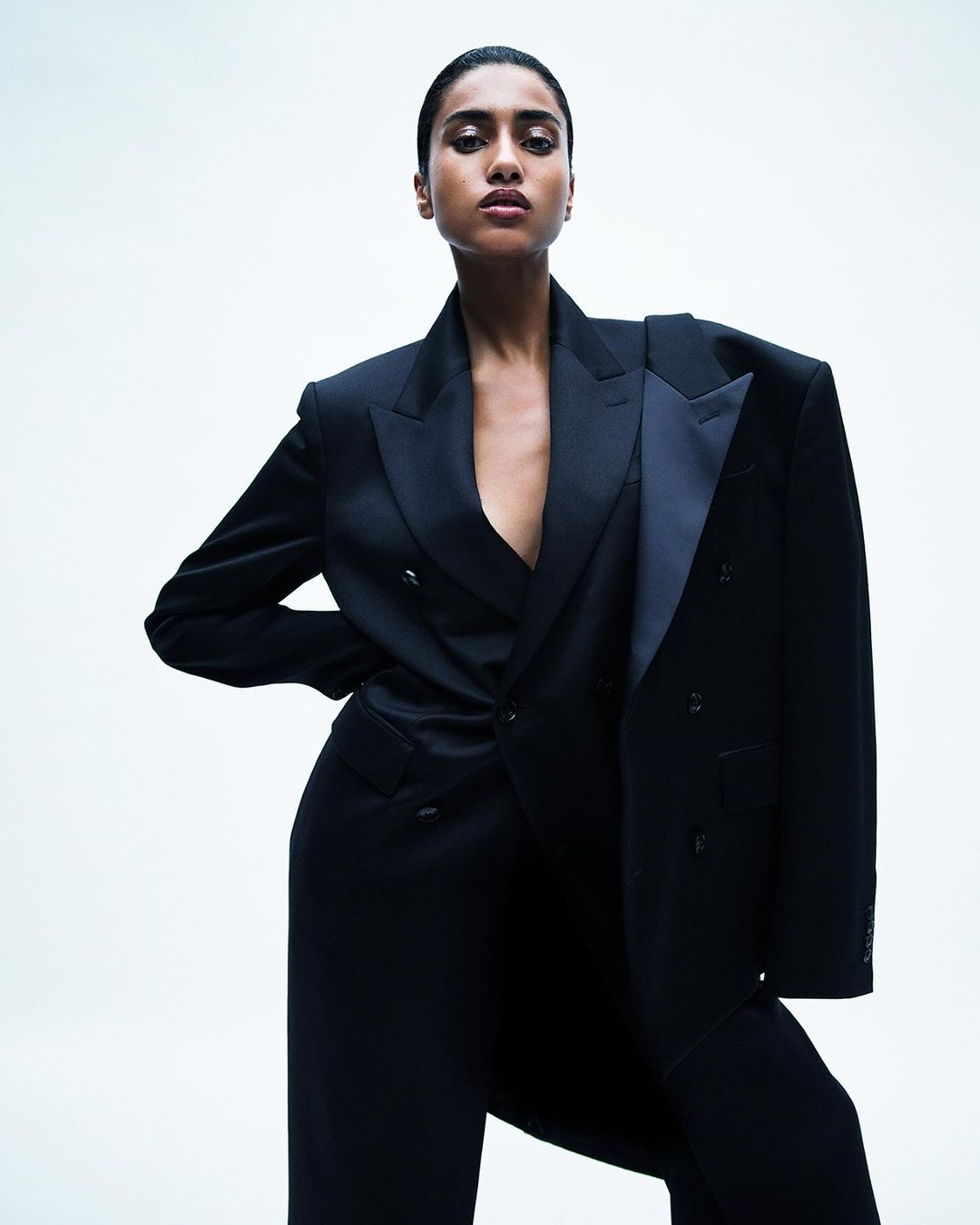 Elegant woman in a black double-breasted suit with an oversized jacket draped over her shoulder against a white backdrop, fashion portrait.