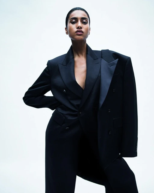 Elegant woman in a black double-breasted suit with an oversized jacket draped over her shoulder against a white backdrop, fashion portrait.