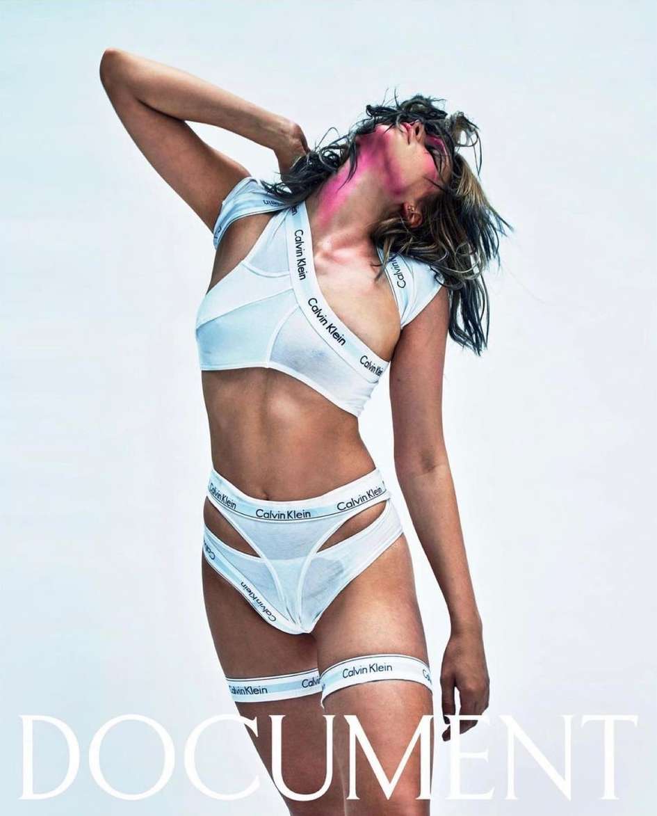 A slender woman poses in a white Calvin Klein crop top and matching underwear set, with pink makeup, against a white background.