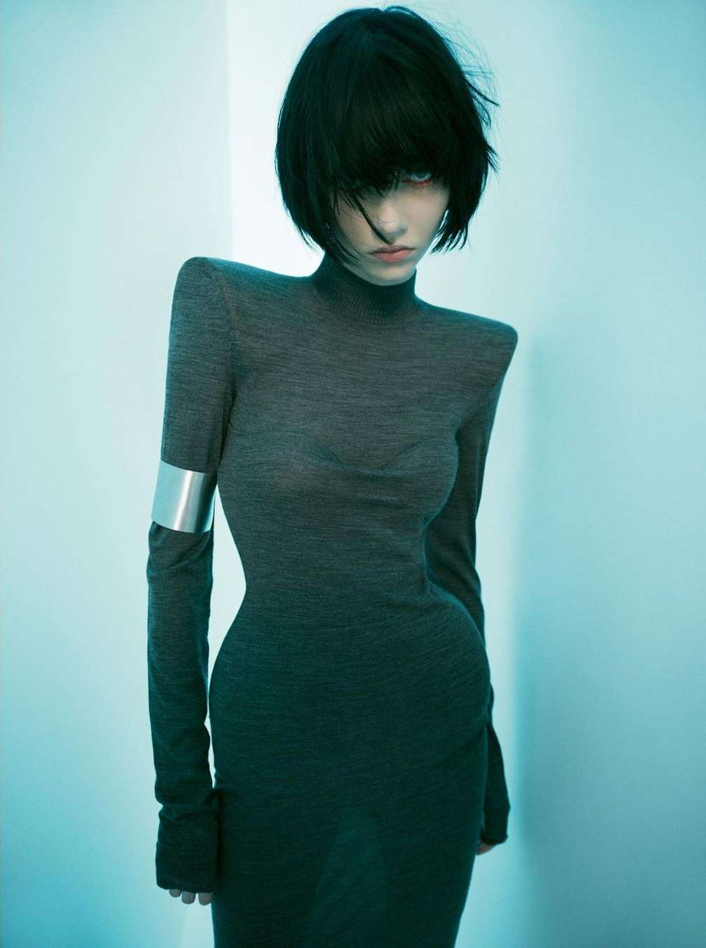 Woman in a long-sleeved gray turtleneck dress poses indoors with soft lighting and black bob haircut; silver cuff on arm.