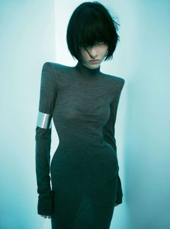 Woman in a long-sleeved gray turtleneck dress poses indoors with soft lighting and black bob haircut; silver cuff on arm.