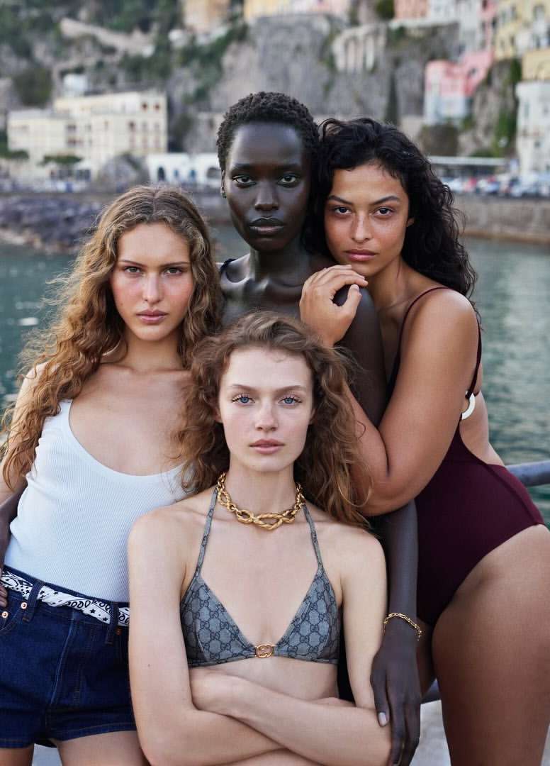 Four diverse women pose in swimwear with a picturesque seaside town in the background, showcasing natural beauty and summer fashion.