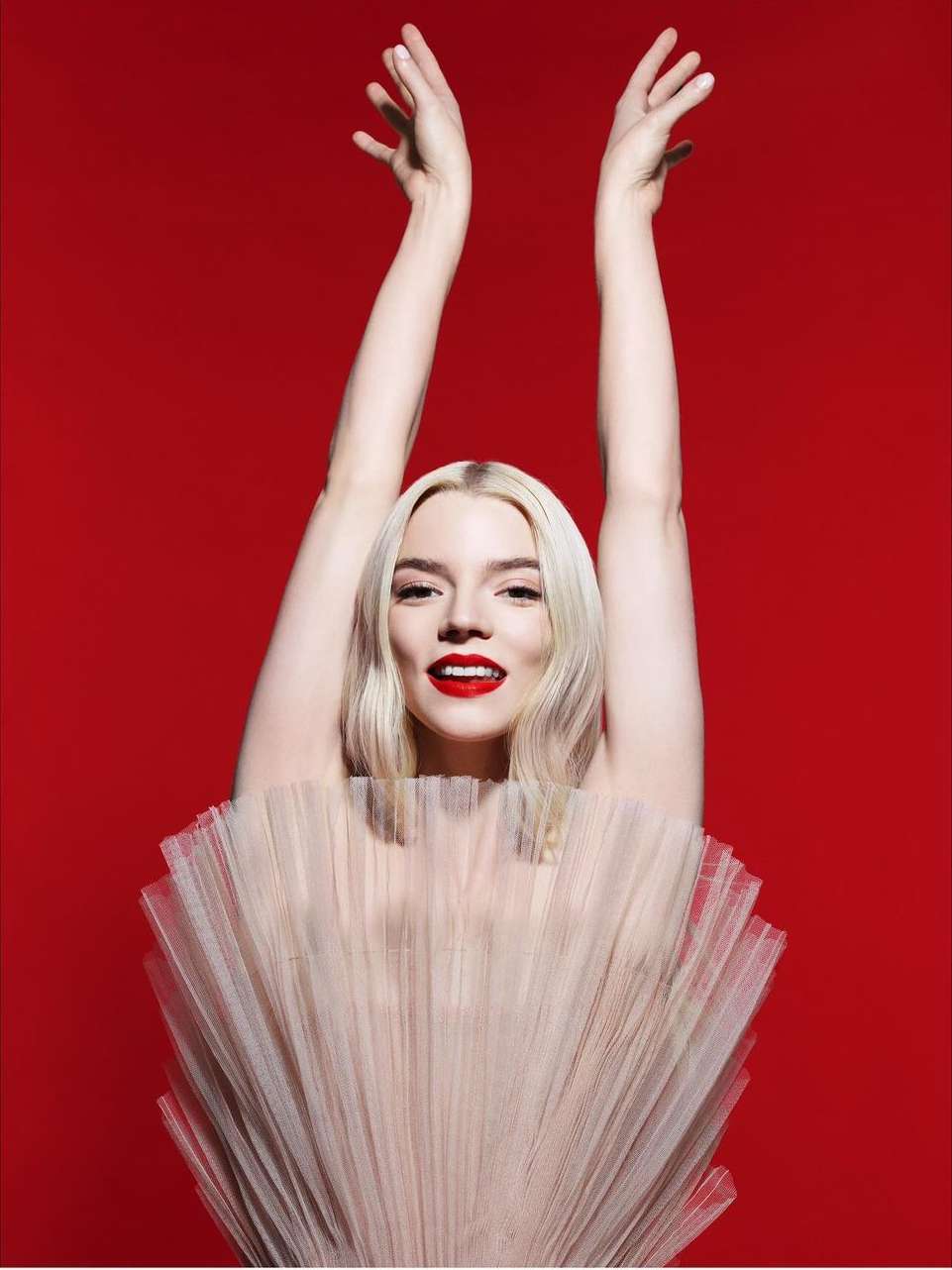 Anya Taylor-Joy poses in a tutu with raised hands against a vibrant red backdrop, showcasing her elegance and poise with a bright red lipstick.