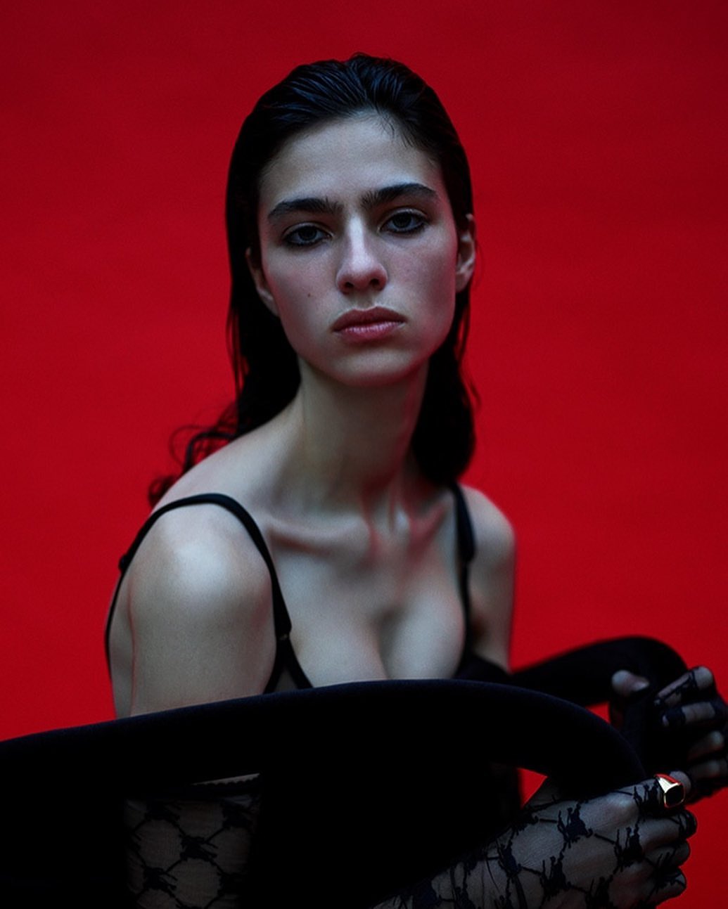 Portrait of an adult woman with dark hair wearing a bra top and lace gloves, posed against a solid red background in a photo studio.