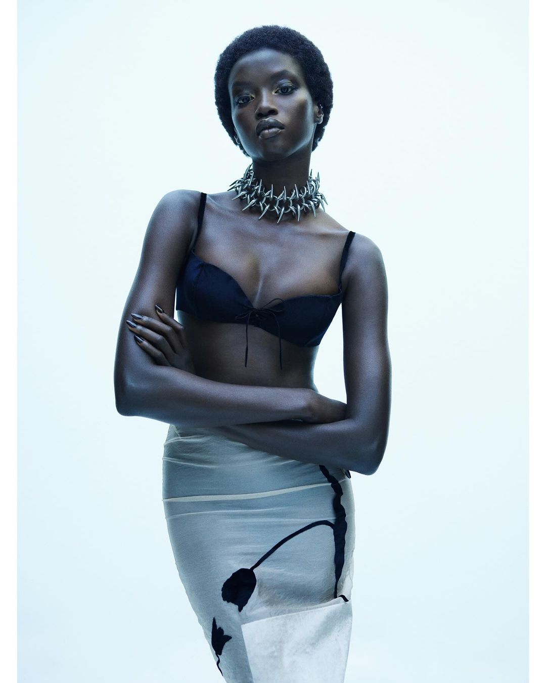 Fashion portrait of a young adult African-descent woman wearing a black bra, spike necklace, and sheer skirt on a white backdrop.