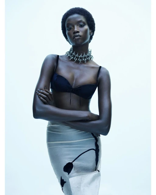 Fashion portrait of a young adult African-descent woman wearing a black bra, spike necklace, and sheer skirt on a white backdrop.