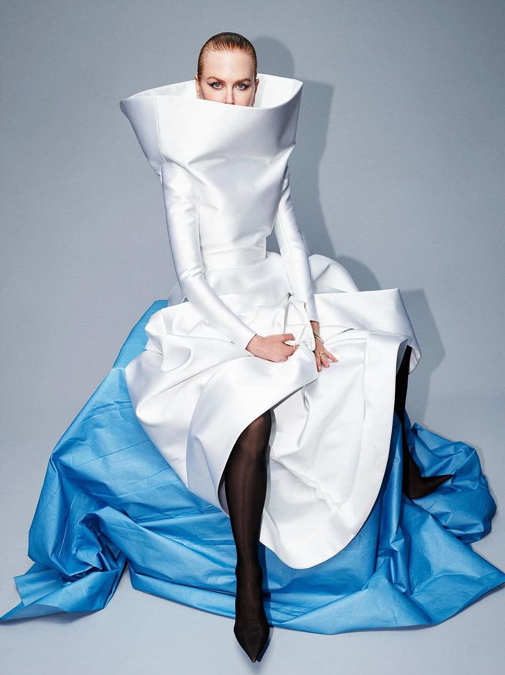 Elegant fashion portrait of a woman in a white dress with a large collar and blue accents, seated against a gray background, modern and stylish.