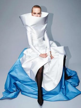 Elegant fashion portrait of a woman in a white dress with a large collar and blue accents, seated against a gray background, modern and stylish.