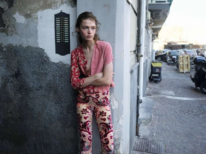 Fashion model in printed pants and jacket poses against a textured grey wall on a city street with parked cars and scooters.