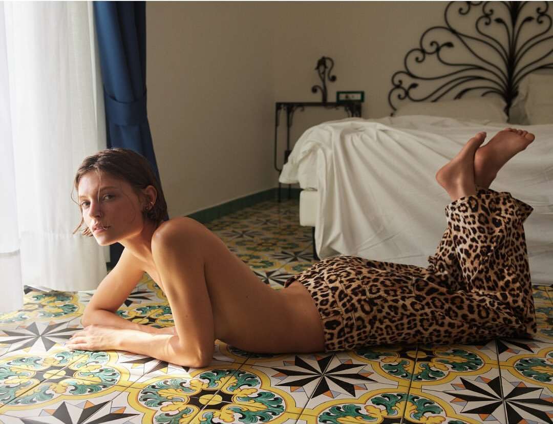 Adult woman lounges on a patterned tile floor wearing leopard print pants near a window with a bed in the background.