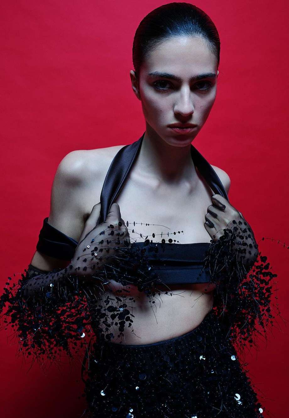 A fair-skinned woman with dark hair poses in a black halter top and skirt with sheer black arm coverings against a red backdrop.