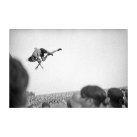Black and white photograph of two people performing an acrobatic move in the air at an outdoor venue with a large crowd in the background.