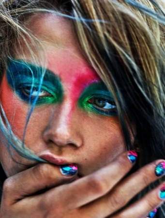 Close-up portrait of a woman with vibrant blue, green, and pink face paint and matching painted fingernails photographed by Mario Sorrenti.