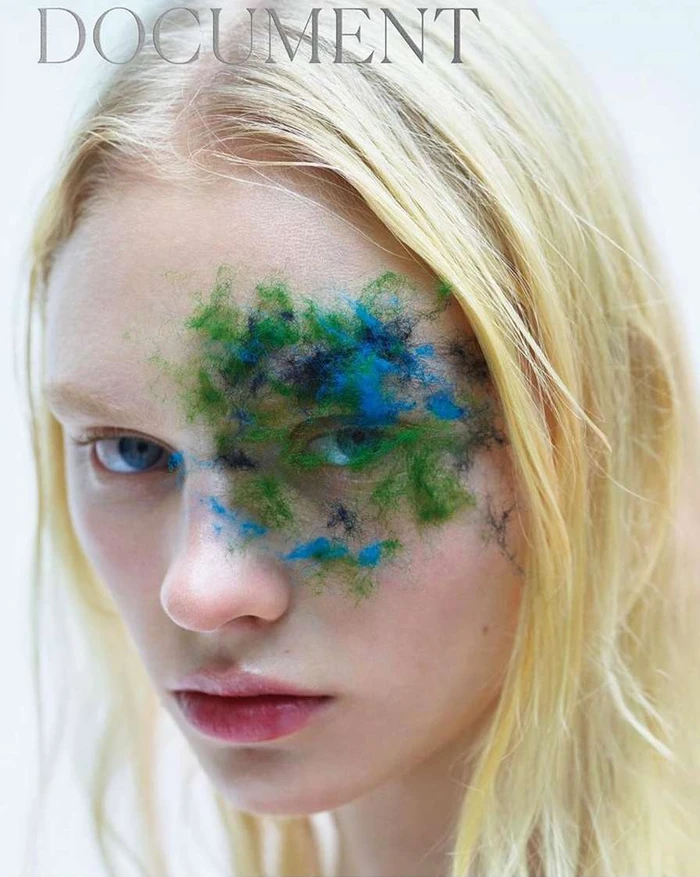 Close-up portrait of a blonde woman with blue eyes and moss or textured green pigment applied over her right eye and cheek.