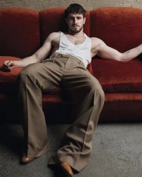 Man reclining on a rust-colored velvet sofa wearing a white tank top, brown trousers, and leather dress shoes