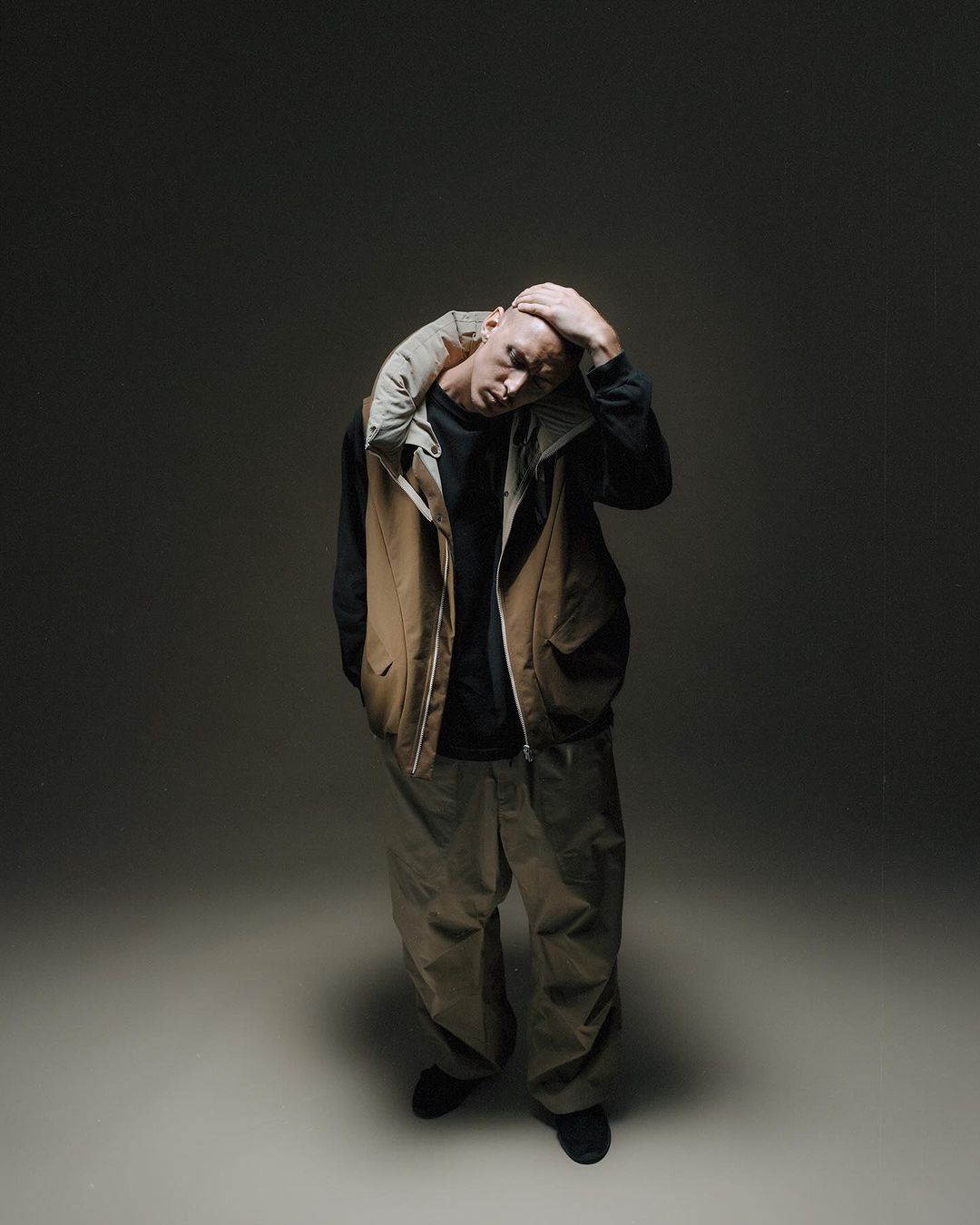 Full body portrait of a bald adult male in casual clothing in a photo studio. The man touches his head and looks down in front of a grey backdrop.