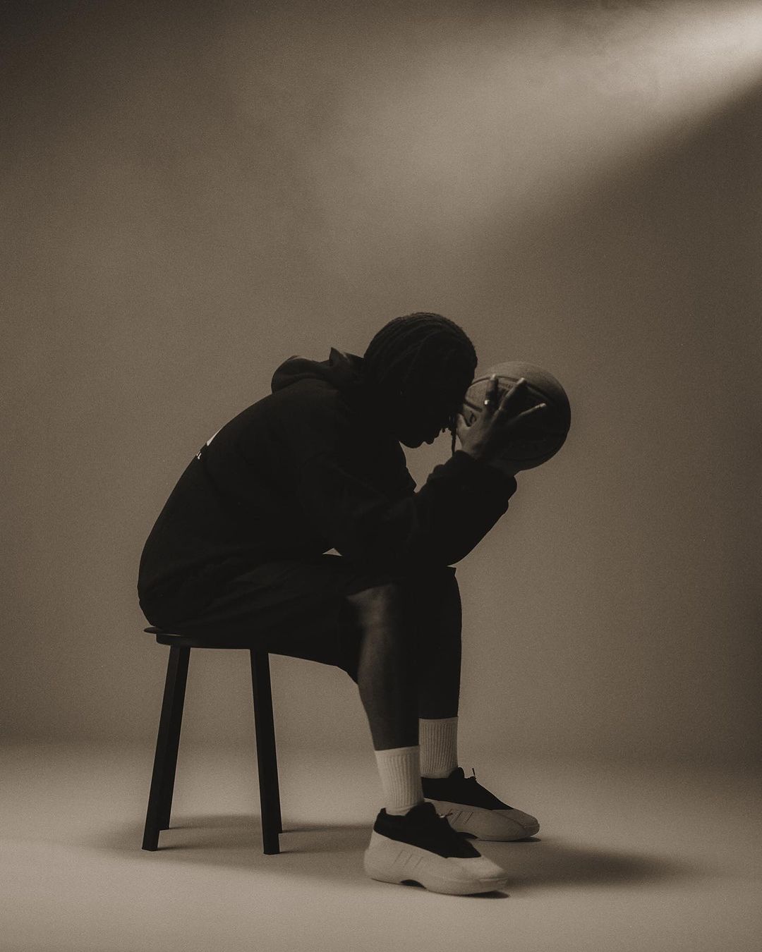 Person in dark hoodie sitting on stool holding basketball under spotlight, wearing sneakers in a neutral-toned studio.
