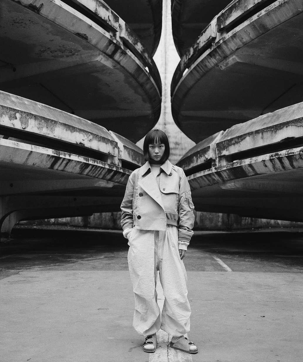 Monochrome portrait of a stylish young woman in a jacket and pants, posing in front of a large Brutalist concrete structure with symmetrical design.
