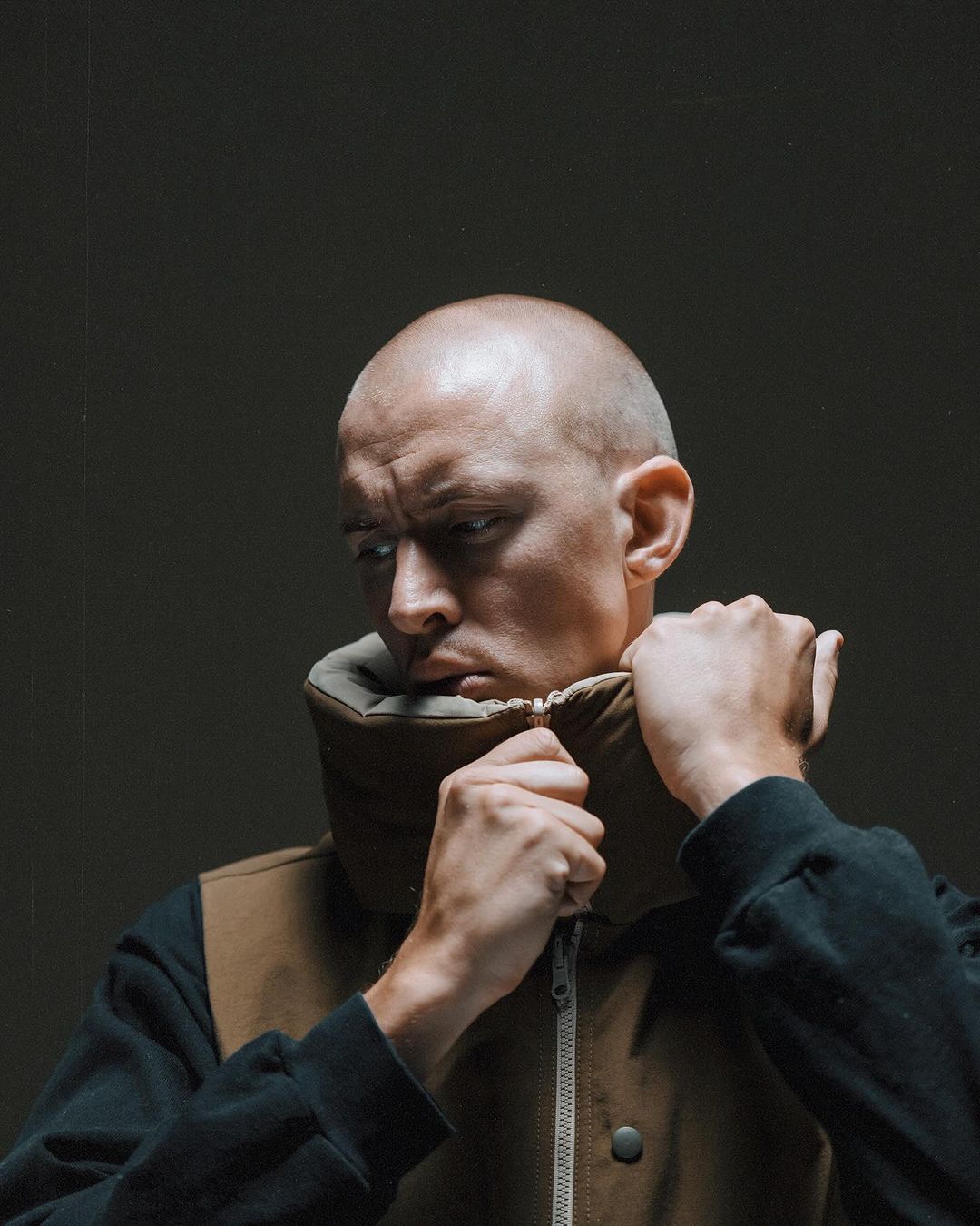 Close-up of a bald man with hands near his neck, wearing a brown vest and a black shirt, against a dark grey background with soft lighting.