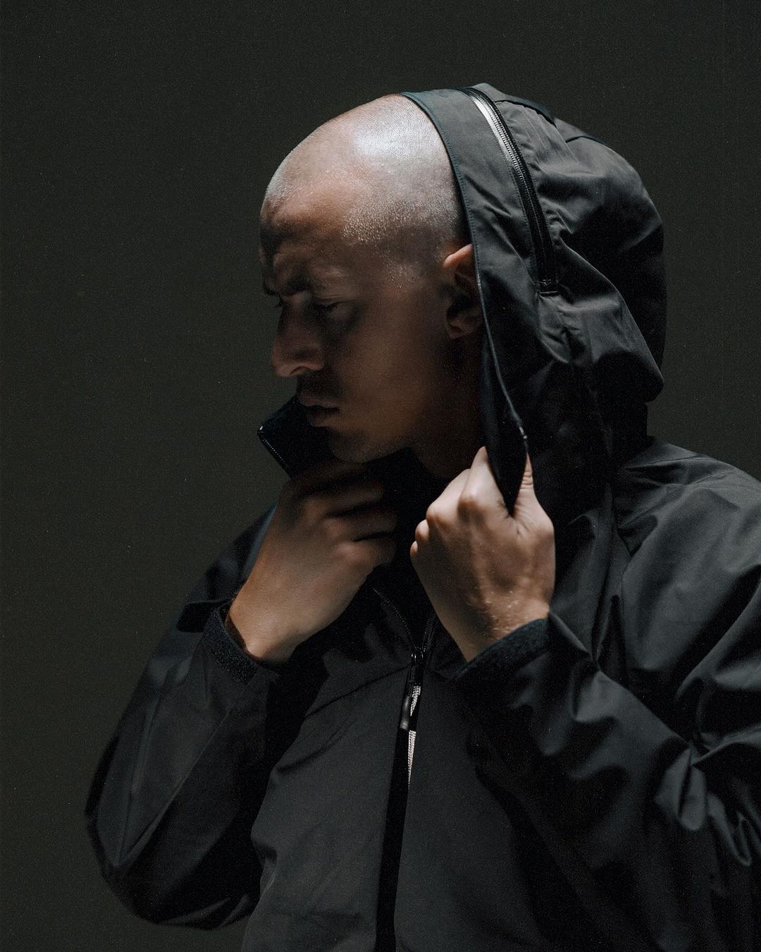 Man in a hooded black jacket zipping up the zipper, shot in front of dark studio background with diffused lighting and a shallow depth of field.