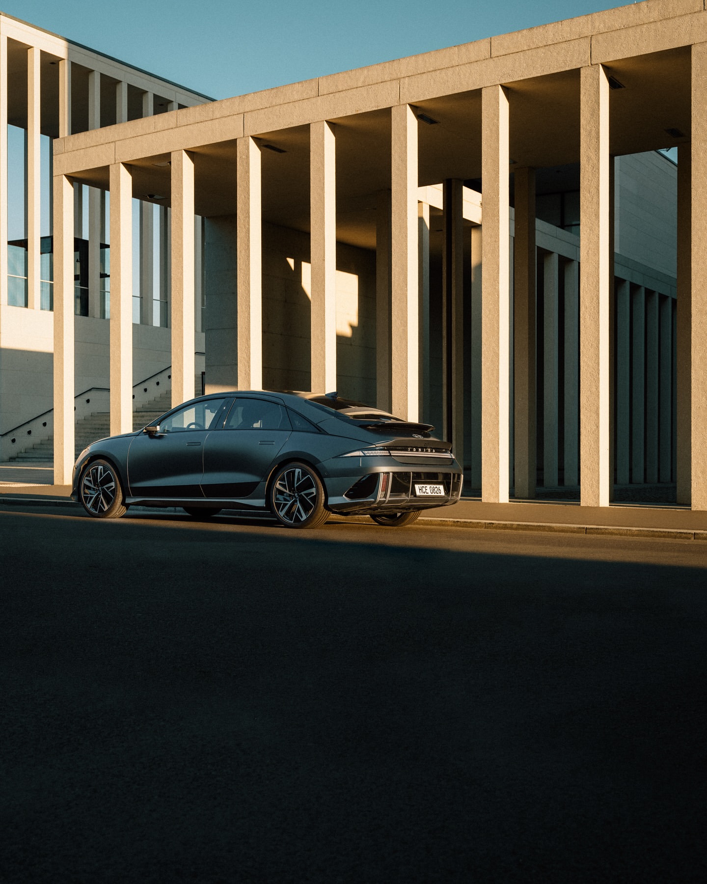 Dark gray Hyundai Ioniq 6 electric car parked in front of a modern building with beige columns on a sunny day. Automotive photography.