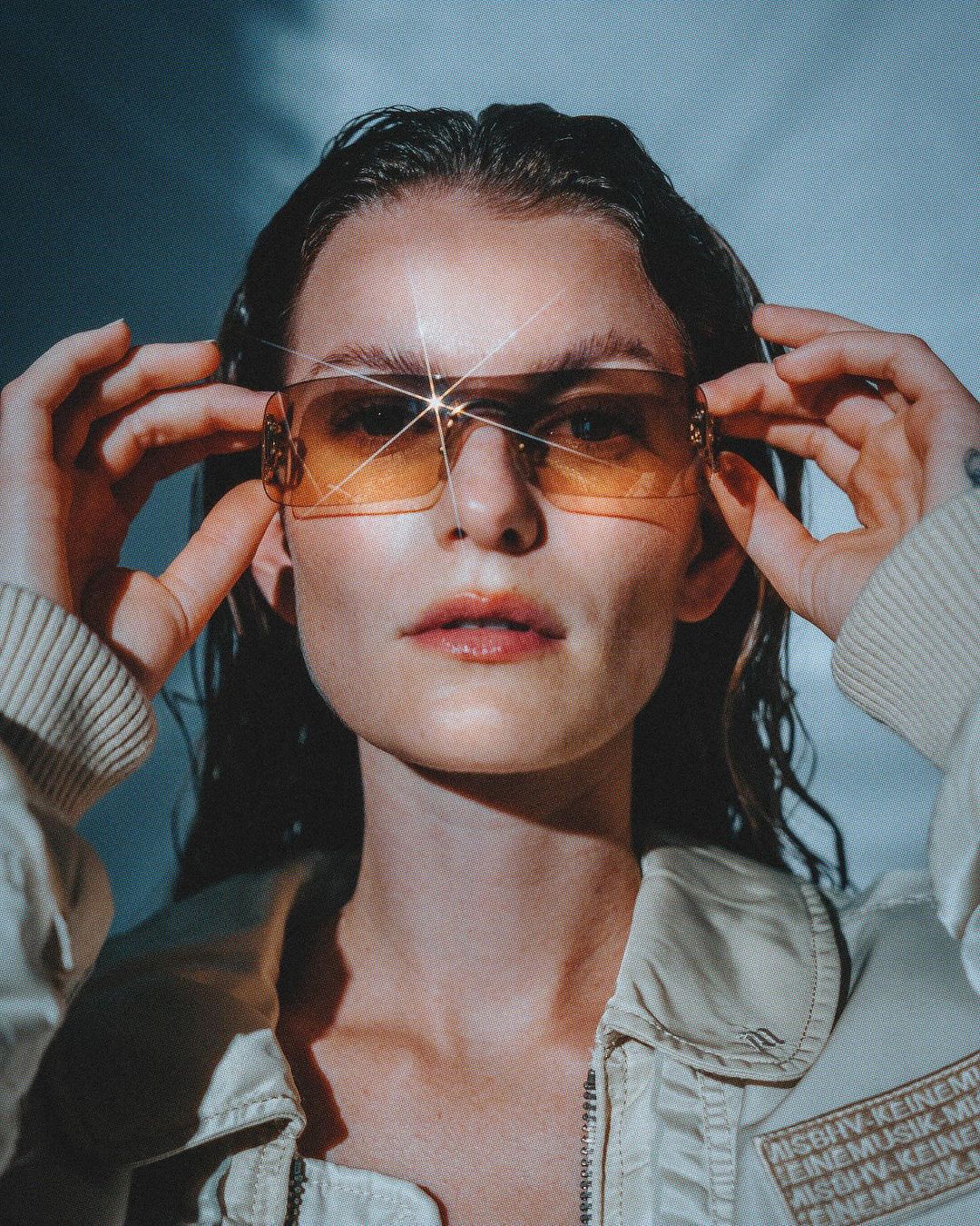 Close-up shot of a young adult woman with fair skin wearing yellow-tinted sunglasses and a light-colored jacket against a blue background.