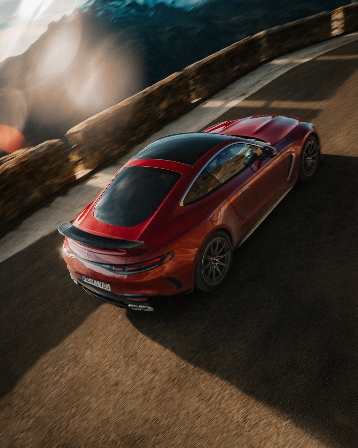 High-angle shot of a red sports car driving on a paved road with a rock wall and blurred mountains in the background, captured during daylight.