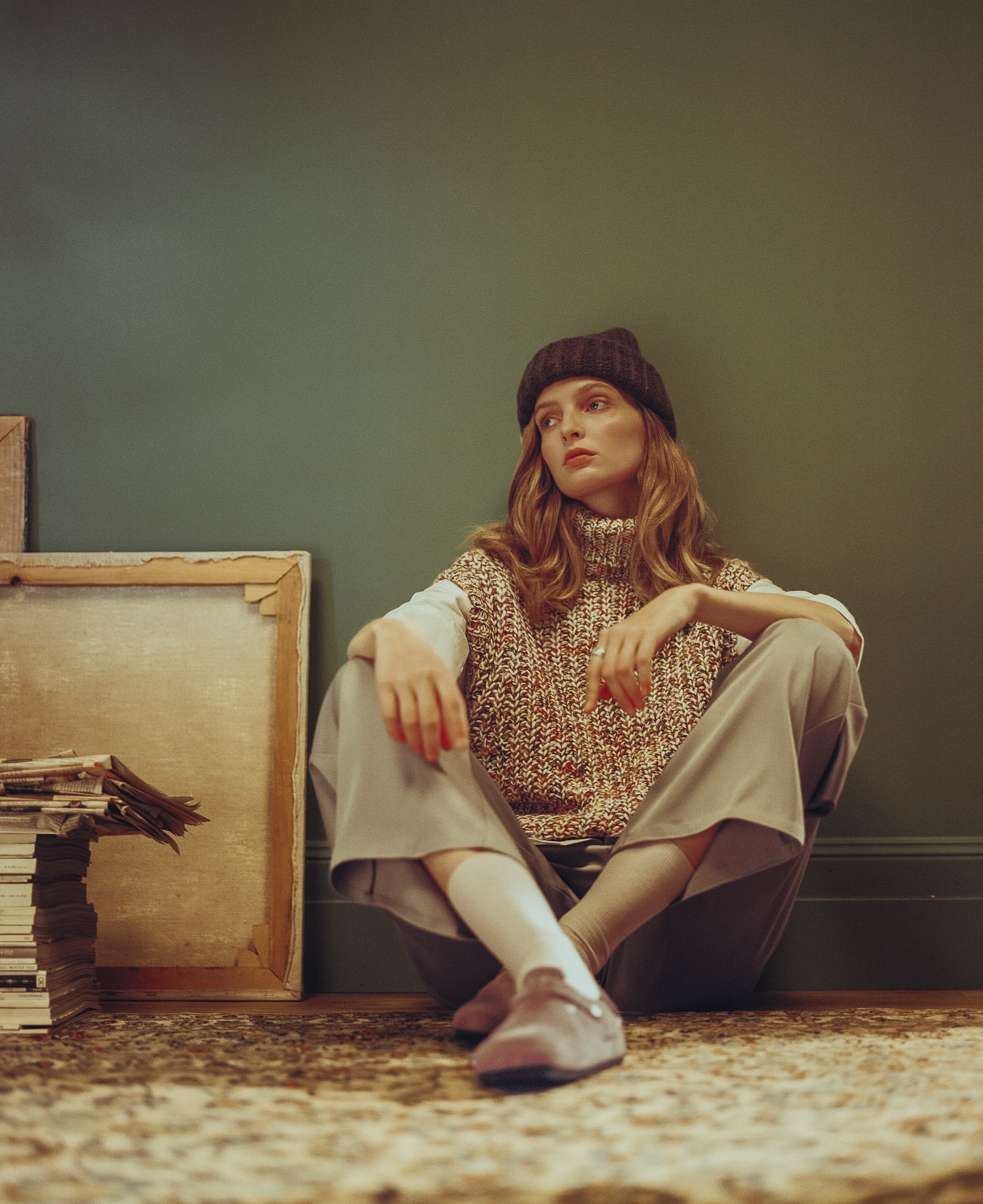 Indoor portrait of a young woman sitting cross-legged against a green wall, wearing a beanie and a patterned sweater vest.
