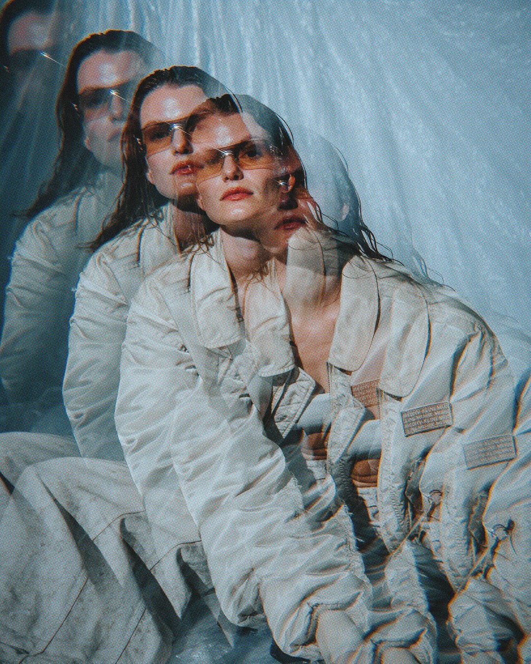A female fashion model poses in sunglasses and jacket with a multiple exposure effect, set against a soft blue background in a studio.