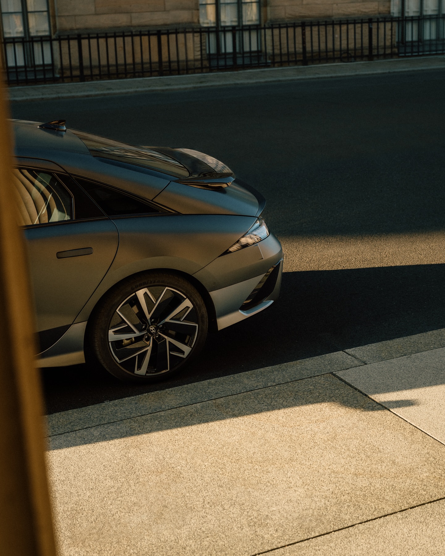 Grey sedan parked on a city street with sunlight reflecting off the vehicle's sleek surfaces, near a building and metal fence.