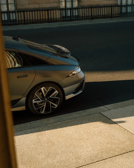 Grey sedan parked on a city street with sunlight reflecting off the vehicle's sleek surfaces, near a building and metal fence.