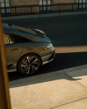Grey sedan parked on a city street with sunlight reflecting off the vehicle's sleek surfaces, near a building and metal fence.