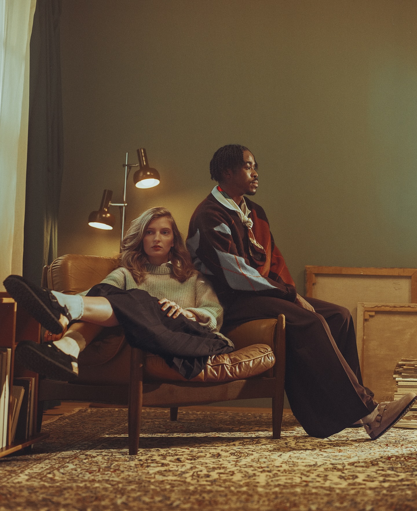 Two people sit in a brown leather chair, one fair-skinned woman with blonde hair and one dark-skinned male in a patterned jacket.
