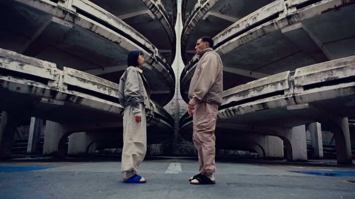 Two people stand face to face on a rooftop under a unique concrete structure with spiral details and faded gray and blue tones.