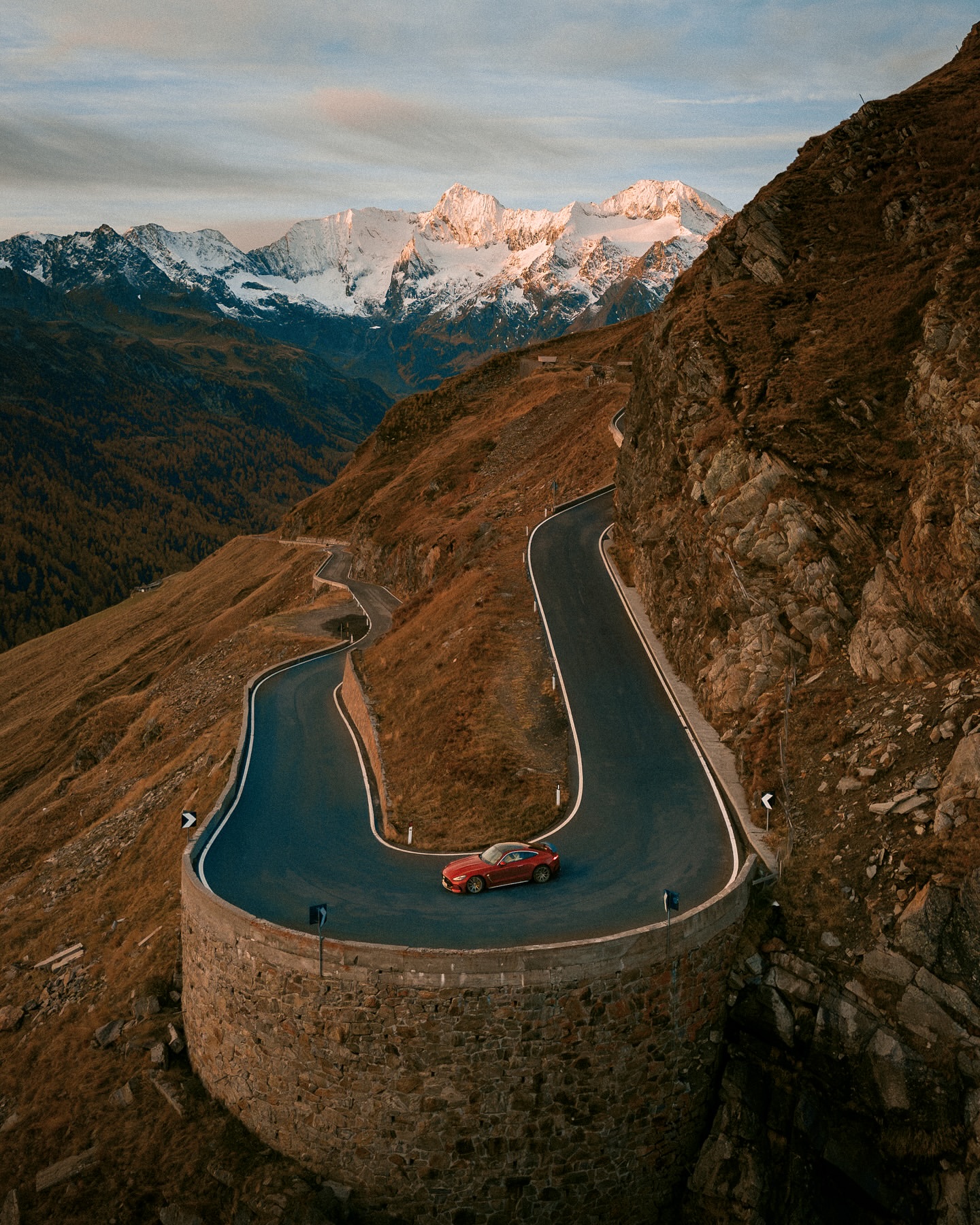 Red sports car drives along a winding mountain road with snow-capped peaks under a bright, partly cloudy sky at sunset or sunrise.