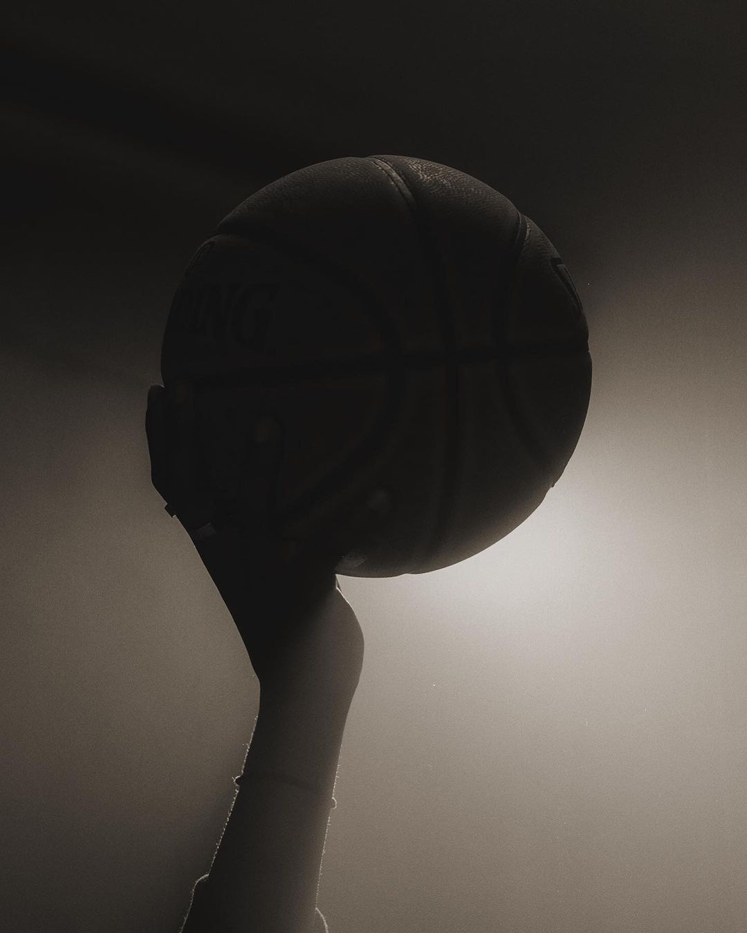 A person holding a basketball on their finger, under a light source, creating a silhouette effect and a moody, monochromatic image.