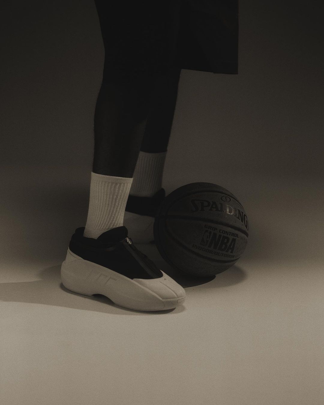 Close-up of a person wearing black and white sneakers and white socks next to a basketball on a grey background in a studio.