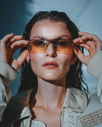 Close-up portrait of a woman wearing tinted orange sunglasses and a light jacket, with a starburst lens flare effect.