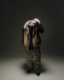 Portrait of a man with shaved head wearing a tan padded vest and khaki trousers against a dark gradient background